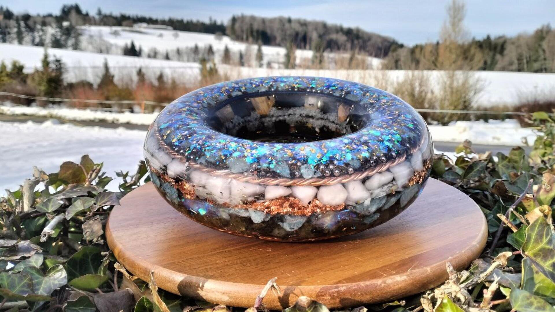 Eine runde, durchscheinende Harzskulptur mit blauen, weißen und kupferfarbenen Akzenten steht auf einem Holzsockel, im Freien mit schneebedeckten Feldern und Bäumen im Hintergrund.