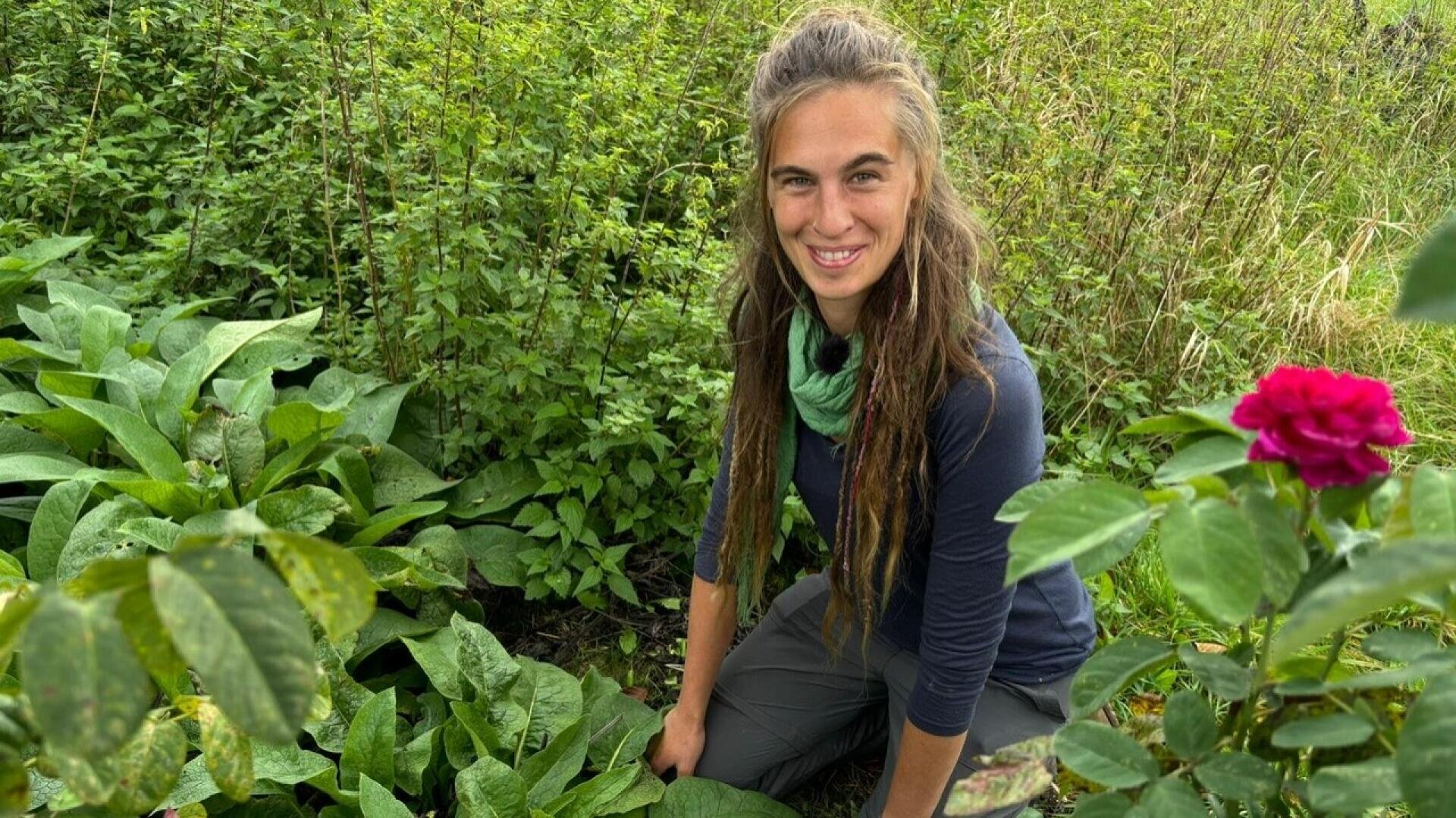 Eine Frau mit langen braunen Haaren und einem grünen Schal kniet in einem üppigen Garten, umgeben von grünen Pflanzen und einer blühenden rosa Rose, und lächelt in die Kamera.