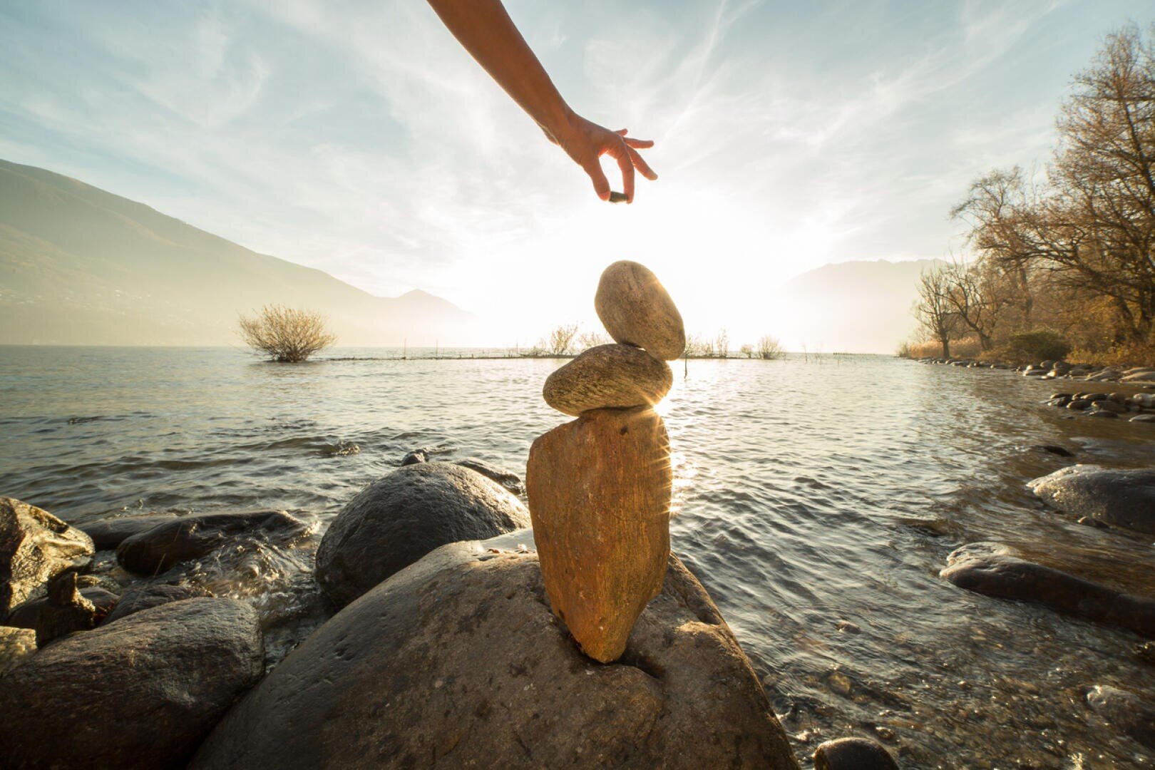 Eine Hand balanciert vorsichtig einen kleinen Stein auf einem Felsenstapel an einem Seeufer bei Sonnenuntergang, mit ruhigem Wasser, fernen Bäumen und Bergen im Hintergrund.