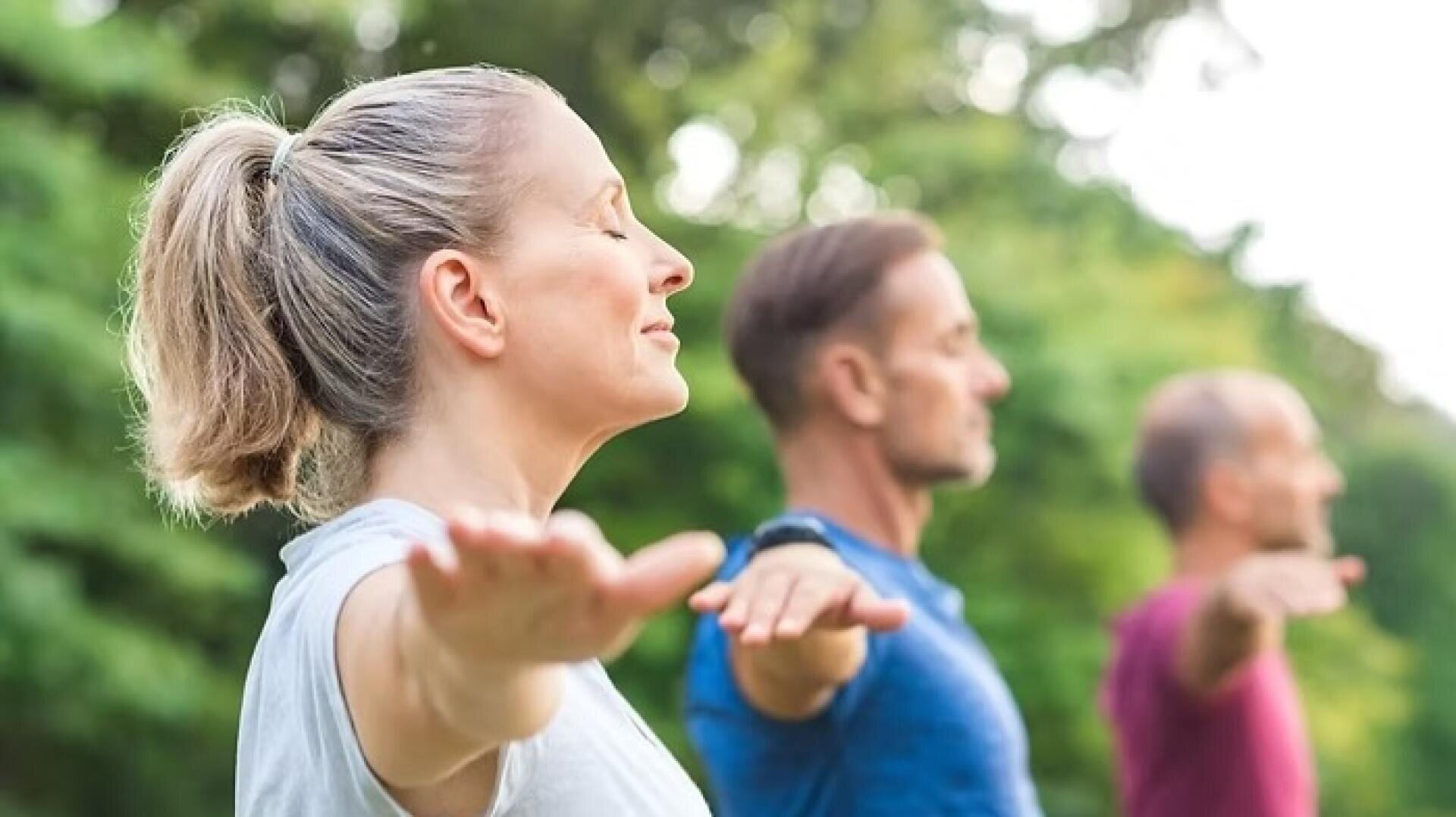 Drei Erwachsene stehen im Freien mit ausgestreckten Armen, geschlossenen Augen und entspannten Gesichtern und praktizieren eine Achtsamkeits- oder Yogaübung im Grünen an einem sonnigen Tag.