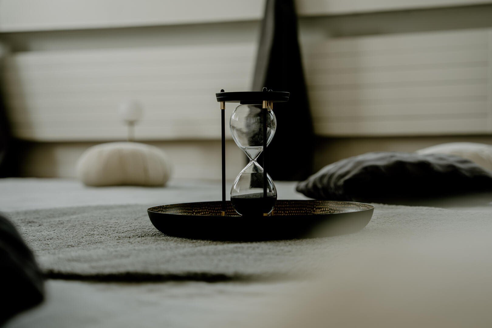 Auf einem grauen Untergrund steht ein dekoratives Tablett, auf dem eine Sanduhr aus Glas liegt.