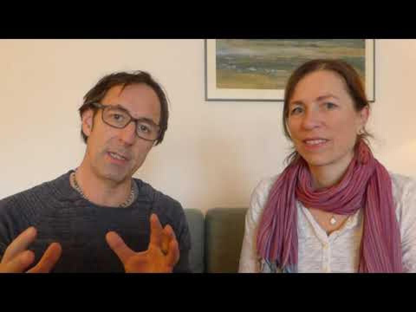 Ein Mann mit Brille und dunklem Pullover gestikuliert beim Sprechen, während er neben einer Frau mit rosa Schal und hellem Hemd sitzt. Beide sitzen in einem Raum auf einer Couch, hinter ihnen hängt ein gerahmtes Bild an der Wand.
