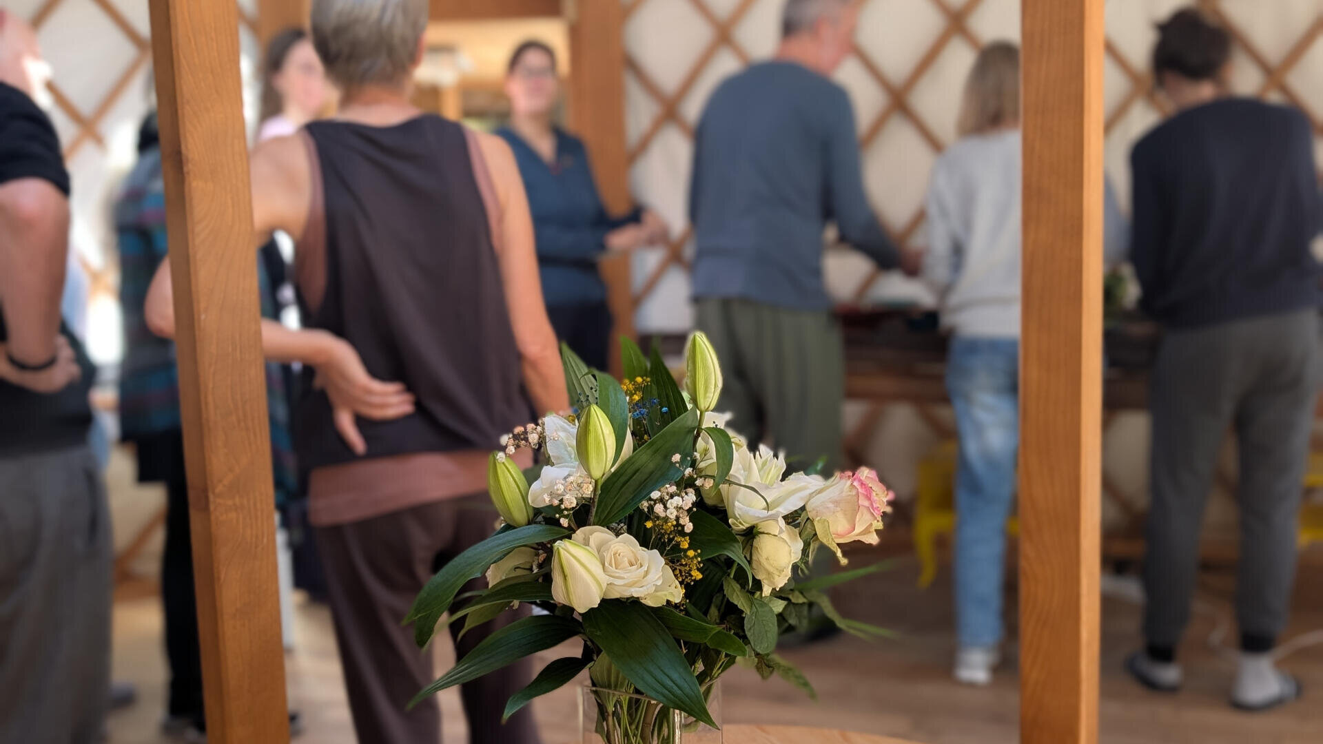 Eine Vase mit weißen und zartrosa Blumen und Grünzeug steht auf einem runden Tisch neben zwei kleinen Zierkürbissen, während sich im Hintergrund eine Gruppe von Menschen in einer Holzjurte unterhält.