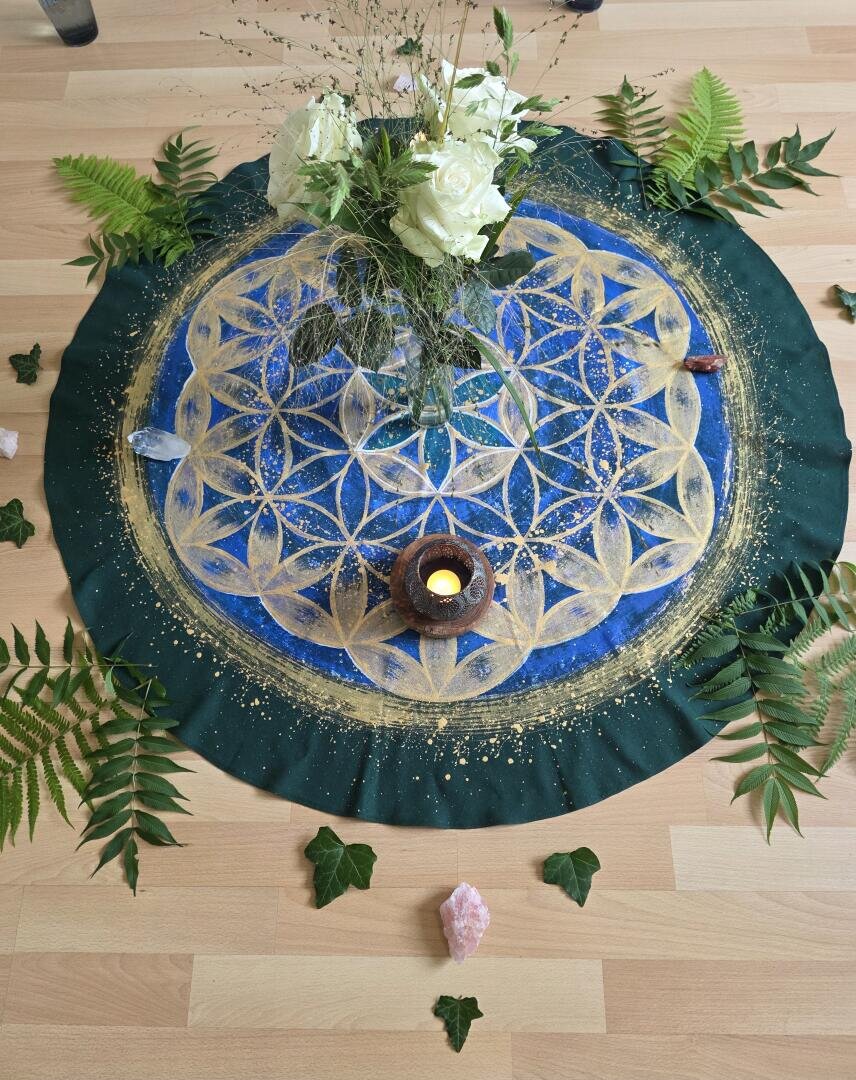 Ein blau-goldenes Mandala-Tuch mit einer brennenden Kerze, weißen Blumen, Kristallen und grünen Blättern, die symmetrisch auf einem Holzboden angeordnet sind.