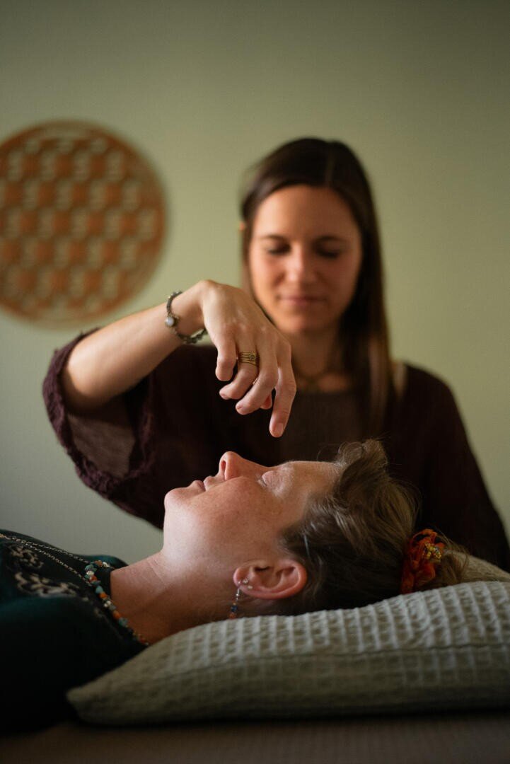 Eine Frau liegt auf einem Kissen, während eine andere Frau ihre Hand über das Gesicht der ersten Frau hält, was auf eine Heil- oder Entspannungsübung in einer ruhigen Umgebung hindeutet.
