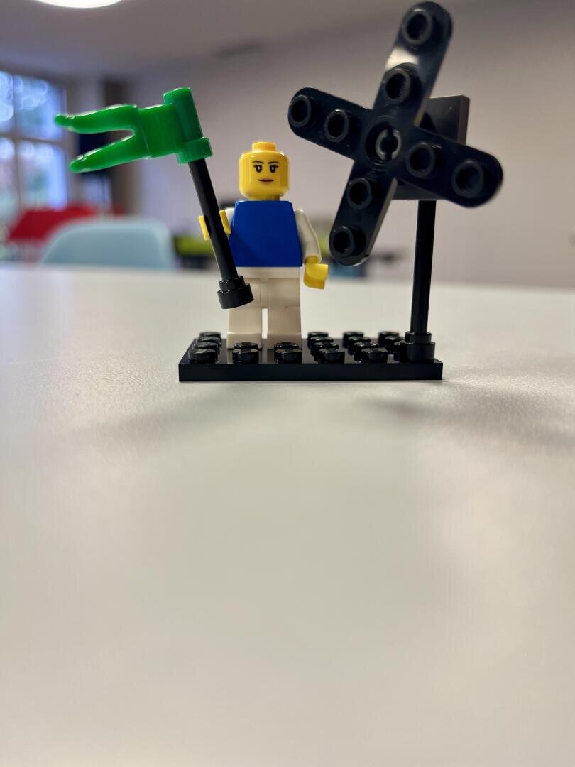 Eine LEGO Minifigur mit einem blauen Hemd und einer weißen Hose steht auf einer schwarzen Grundplatte. Die Figur hält eine grüne Flagge in der Hand, daneben steht ein schwarzes windmühlenähnliches Gebilde. Der Hintergrund ist eine unscharfe Innenraumkulisse.