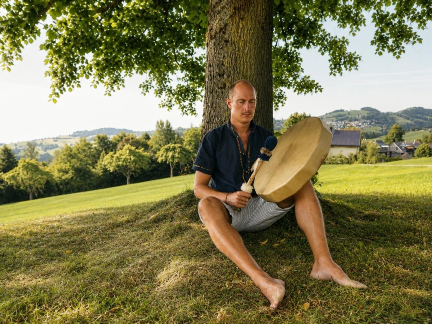 Ein Mann sitzt am Fuße eines Baumes und spielt mit einem Schlägel auf einer Rahmentrommel.