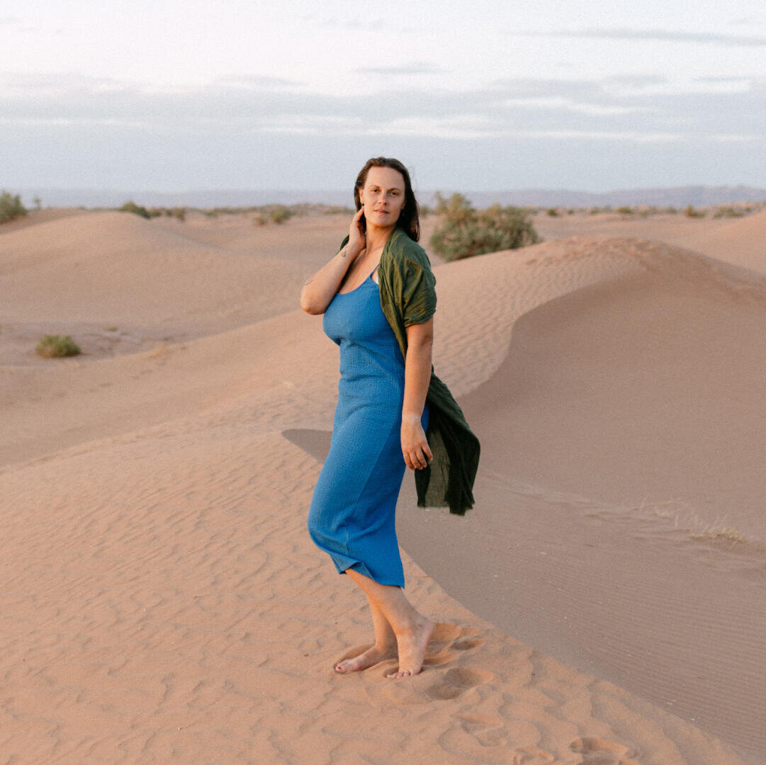 Eine Frau in einem blauen Kleid und einem grünen Schal steht barfuß auf Sanddünen und posiert mit einer Hand nahe ihrem Hals. Im Hintergrund sind weitere Dünen und ein teilweise bewölkter Himmel zu sehen.