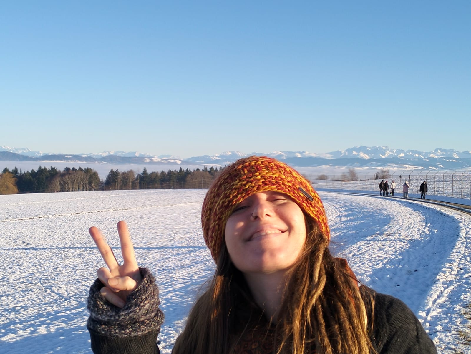 Eine lächelnde Person, die eine bunte Strickmütze und Handschuhe trägt, zeigt ein Friedenszeichen in einer verschneiten Landschaft mit klarem blauen Himmel, fernen Bergen und Menschen, die einen verschneiten Weg im Hintergrund entlanggehen.