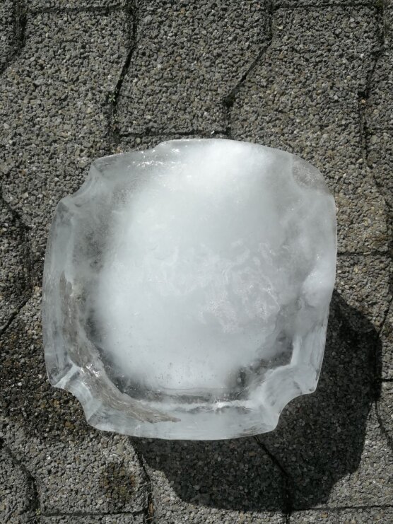 Ein großer, annähernd kreisrunder Eisblock mit unregelmäßigen Kanten liegt auf einem strukturierten, grauen Steinpflaster im Sonnenlicht.
