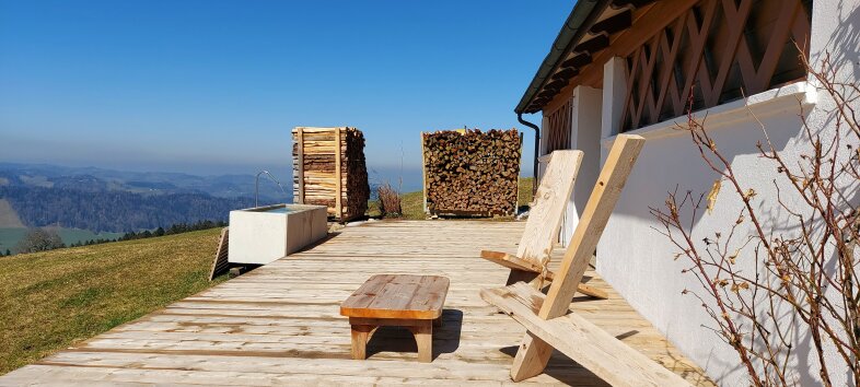 Holzterrasse mit zwei Liegestühlen, einem kleinen Tisch und zwei Stapeln Brennholz, mit Blick auf eine Berglandschaft.