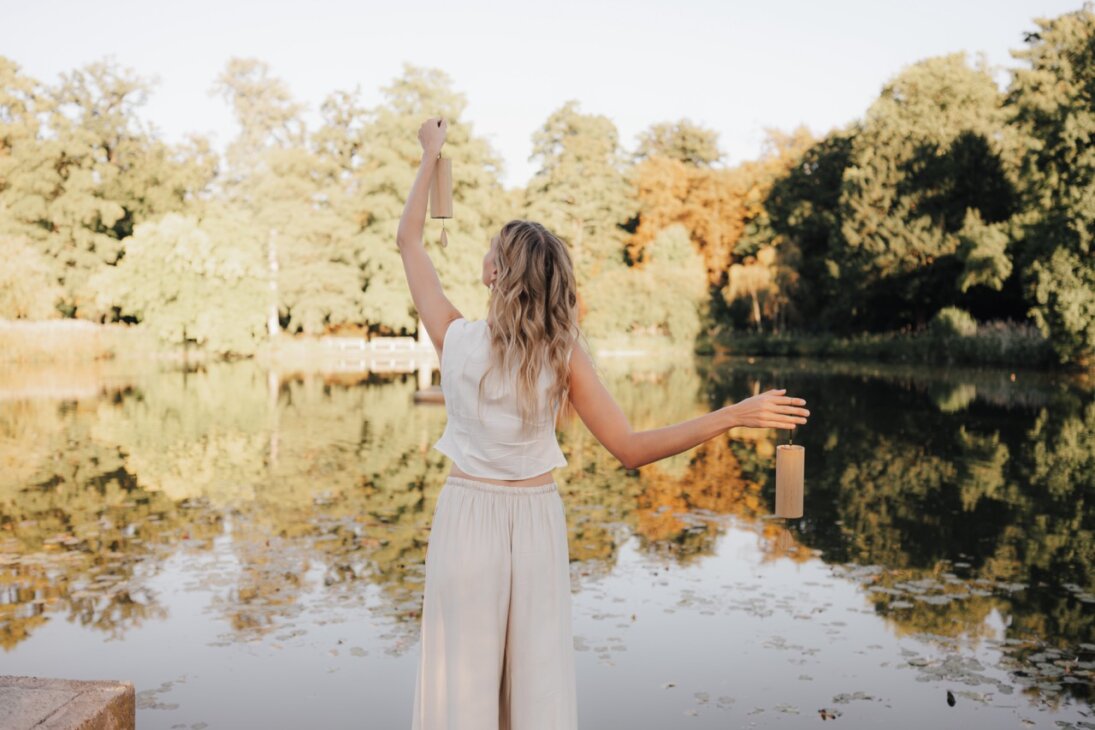 Eine Frau in leichter Kleidung steht an einem ruhigen See und hält mit erhobenen Armen zwei Laternen. Sie blickt von der Kamera weg auf das Wasser und die Bäume unter einem klaren Himmel.